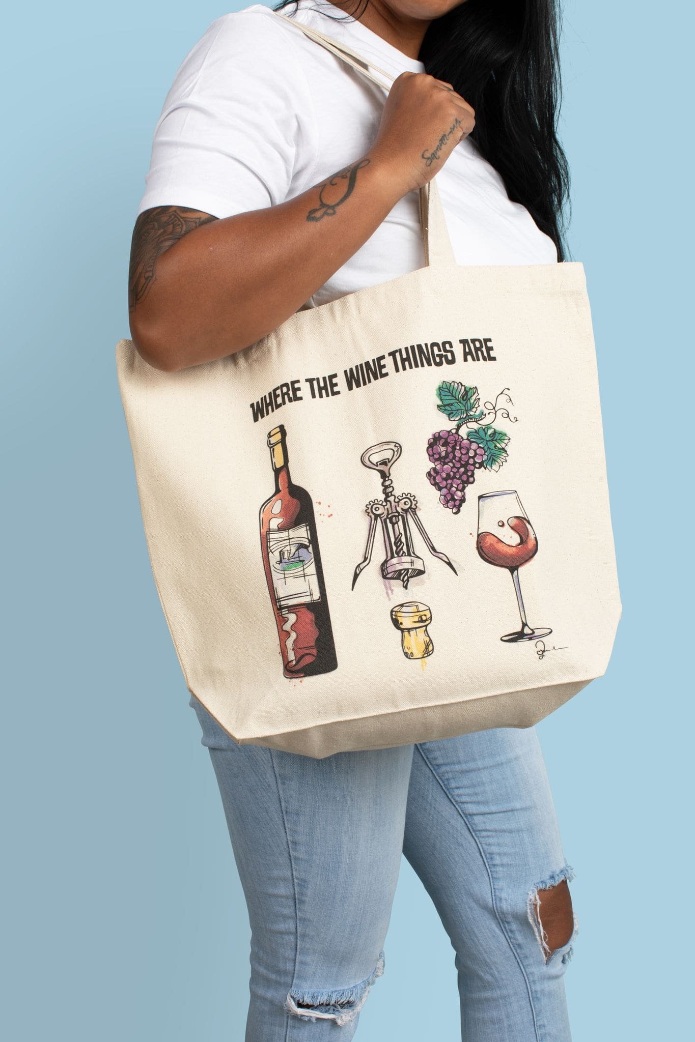 Woman holding canvas hopping tote bag with illustration of wine bottle, grapes, wine glass, cork, and corkscrew