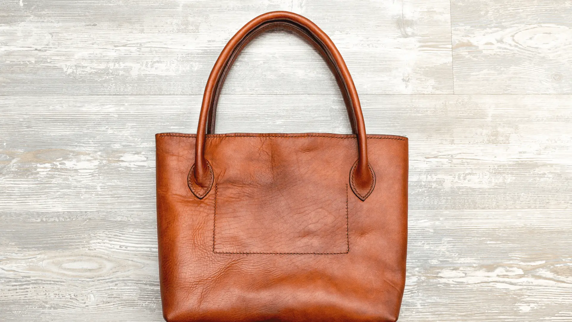 Brown leather tote bag with two handles and a front pocket on a wooden surface.