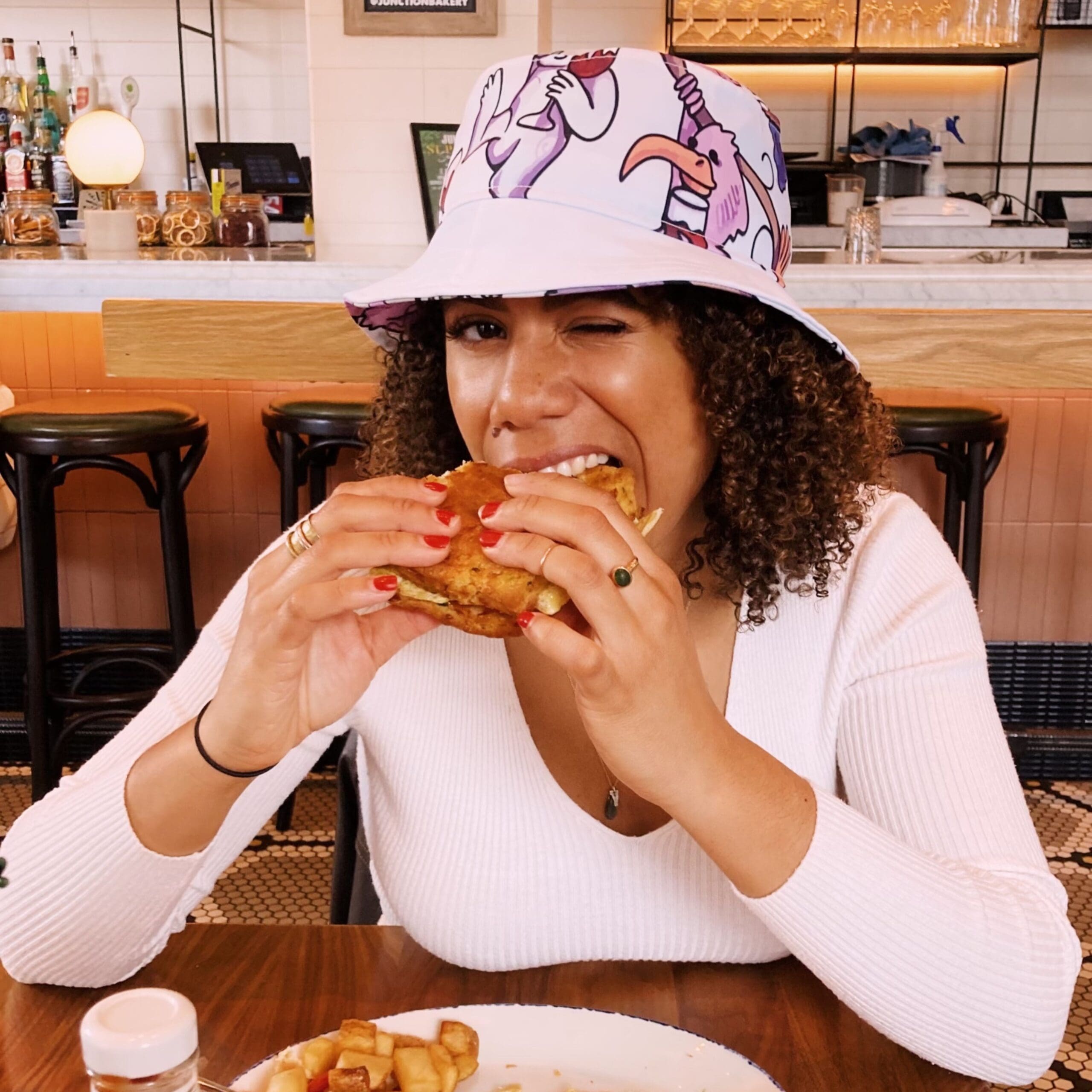 A woman wearing a bucket hat sitting at a table eating a sandwich